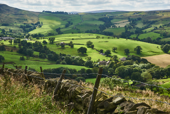 Peak district This landscape photograph shows the rural scenery of the Peak District in the United Kingdom during a summer afternoon. The image features rolling green hills characteristic of the area, separated by traditional stone walls and intermittent lines of trees, with patches of farmland and scattered stone-built houses typical of rural English countryside. The gentle sunlight and partial clouds create areas of contrasting light and shadow across the fields, highlighting the natural beauty and agricultural heritage of the region. The Peak District is renowned for its nature, expansive open land, and picturesque settings, making it a significant destination in the United Kingdom.